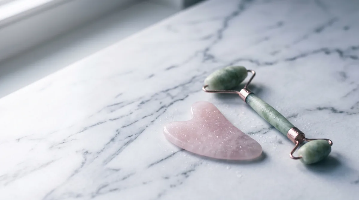 Rose quartz gua sha tool and jade roller on marble surface with condensation droplets in humid climate